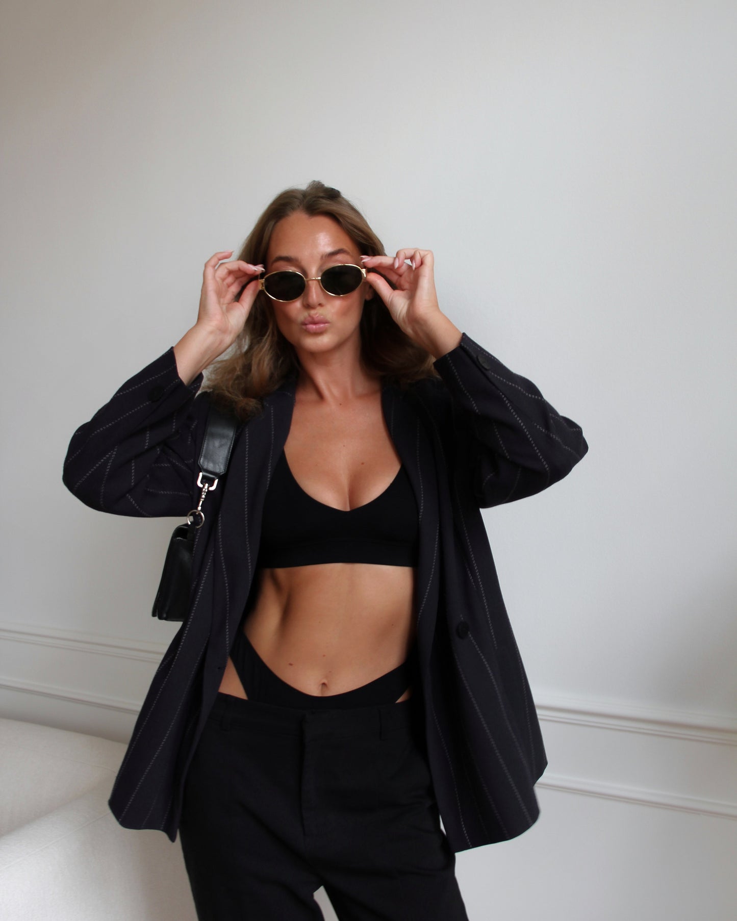 Woman wearing a deep V bralette and high-cut underwear styled with a black blazer, tailored trousers, and sunglasses.