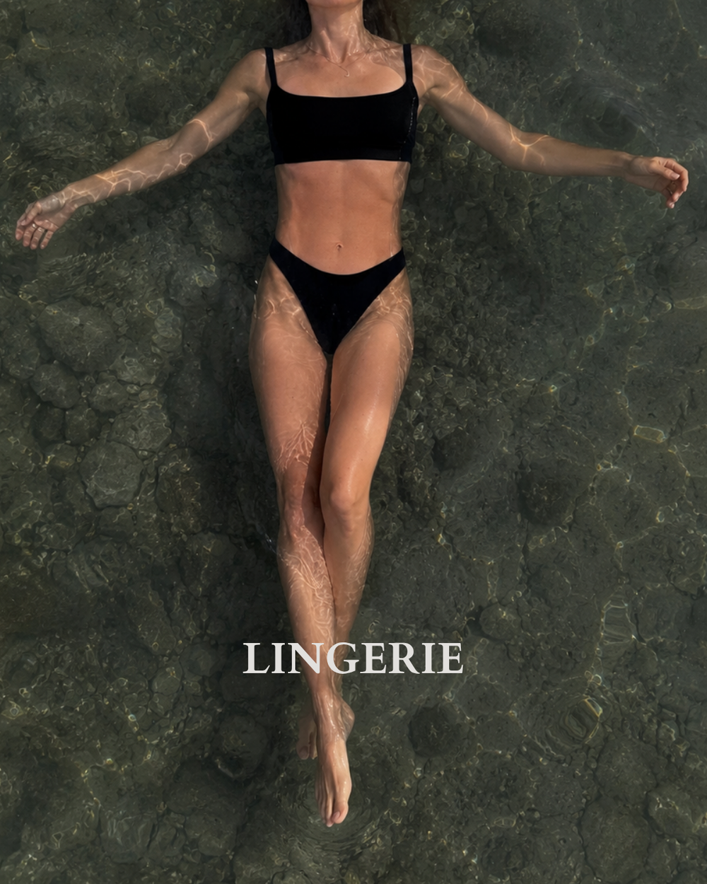 A woman floating in shallow water wearing a black two-piece lingerie set from Baume Essentials, with the word 'LINGERIE' printed in white near the bottom.
