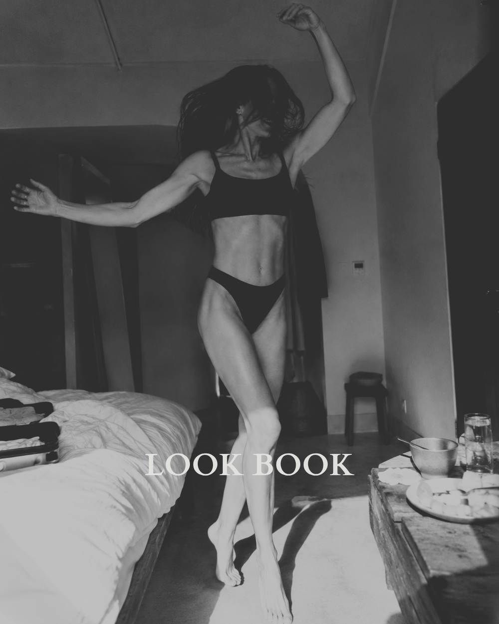 A black and white lifestyle photograph of a woman in a black lingerie set from Baume Essentials posing in a bedroom, with the text 'LOOK BOOK' overlaid at the bottom.