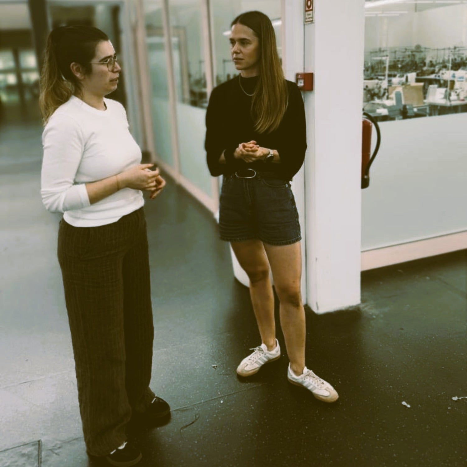 Two women standing and talking in a modern office hallway with glass walls and white columns.