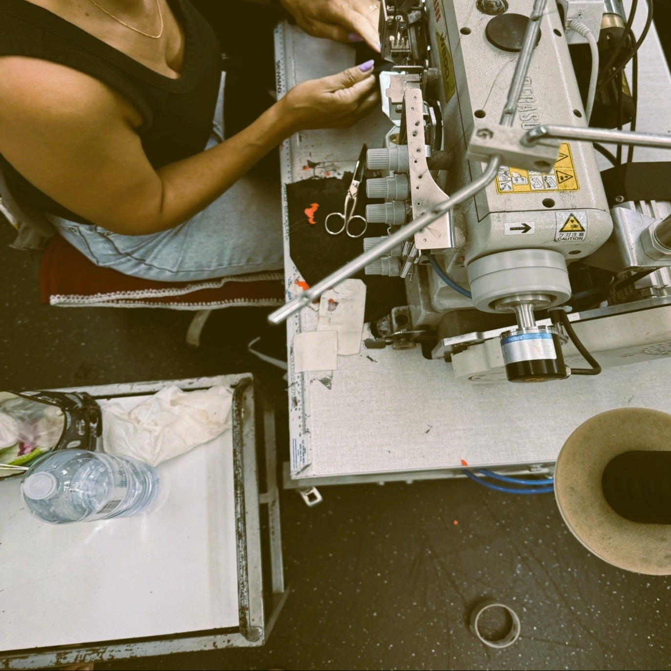 A woman wearing glasses and a black tank top operates a large industrial sewing machine at a worktable in a workshop setting.
