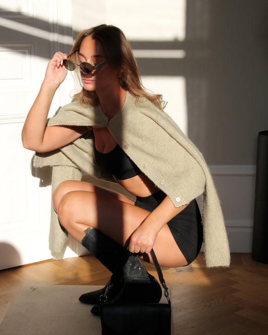 Woman wearing a stylish bralette and shorts set, paired with sunglasses and a knitted cardigan, seated on the floor in a relaxed pose.