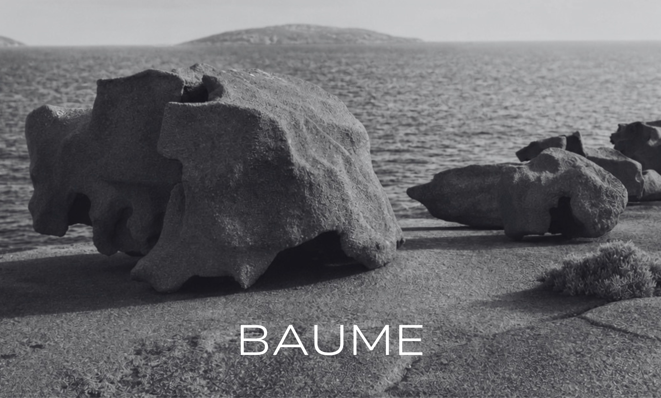 Black and white photograph of large rocks on a beach with 'BAUME' branding.