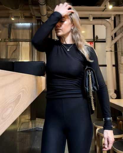 Woman wearing BAUME black leggings and a matching long-sleeve top, styled as an elevated everyday athleisure look in an industrial loft.