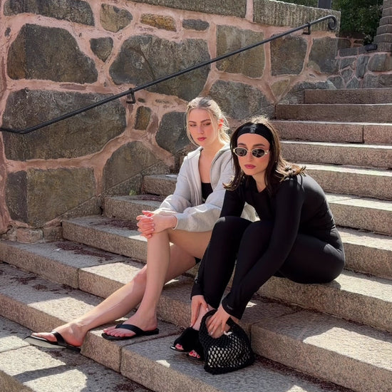 Two women wearing BAUME Form collection pieces on stairs, one in black tights and a long-sleeve top with heels, jewelry and a bag, the other in a black bralette and shorts with a hoodie and flip flops.