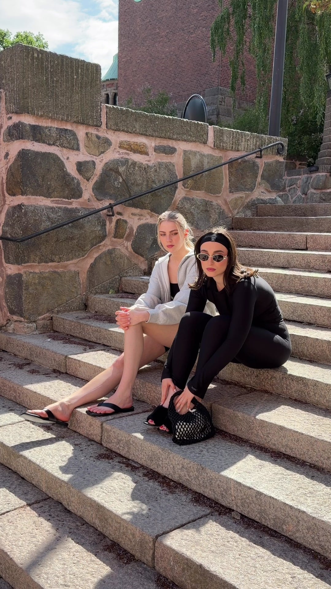 Two women wearing BAUME Form collection pieces on stairs, one in black tights and a long-sleeve top with heels, jewelry and a bag, the other in a black bralette and shorts with a hoodie and flip flops.