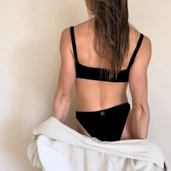 Co-founder Denise from behind wearing a sensual soft lingerie set from Baume Essentials and holding a light gray robe against a plain beige wall.