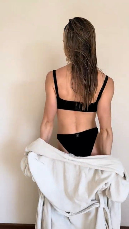 Co-founder Denise from behind wearing a sensual soft lingerie set from Baume Essentials and holding a light gray robe against a plain beige wall.